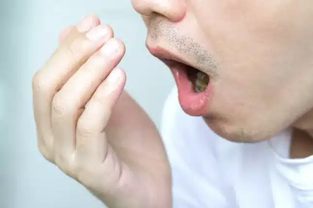 An Asian man holding his hand up to his mouth and nose to check the smell of his breath.